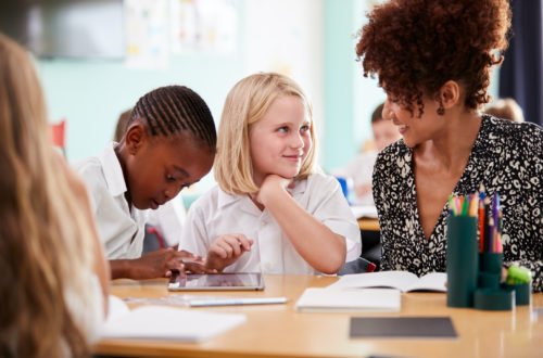 Female Teacher With Two Elementary School Pupils Wearing Uniform Using Digital Tablet At Desk