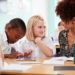 Female Teacher With Two Elementary School Pupils Wearing Uniform Using Digital Tablet At Desk